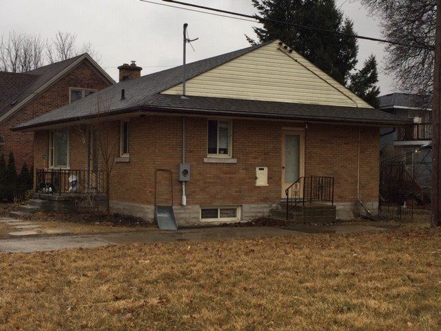 A tan brick bungalow with a grey shingled roof, light siding in the gable, and a small porch on an overcast day.