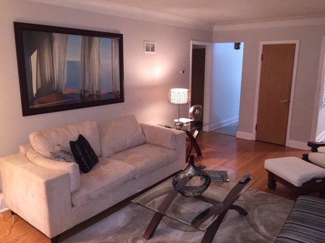 A beige sofa, glass coffee table, and large framed artwork in a sunlit living room with hardwood floors.