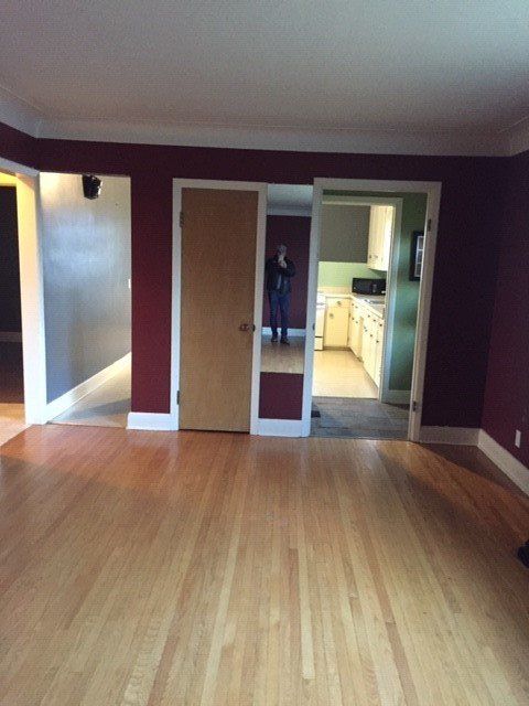 A room with dark red walls and hardwood floors, featuring a wooden door, a mirror, and a view into a kitchen.