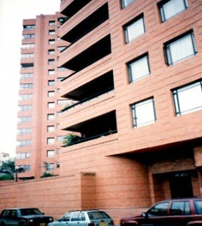 A tall, reddish-brown brick apartment building with recessed balconies and windows, viewed from a street with parked cars.