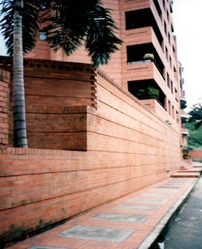 A tall brick building with tiered balconies rises above a long, textured brick wall and a paved sidewalk.