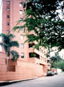 A multi-story, reddish-brown apartment building with balconies, surrounded by green trees on a street with parked cars.