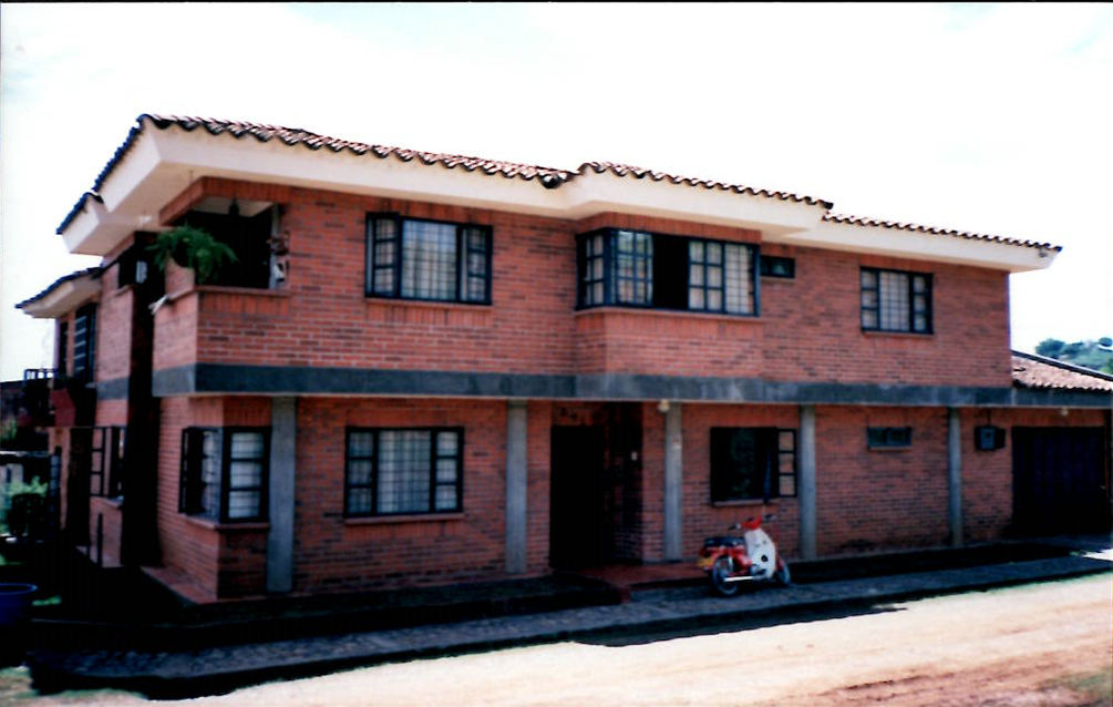 A two-story red brick house with a tiled roof and several windows, featuring a small motorcycle parked in front.