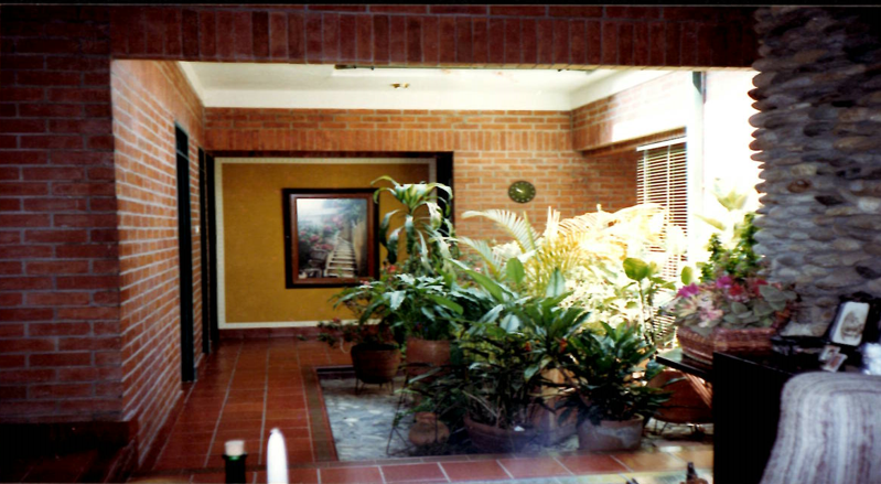 An indoor courtyard with red brick walls, a tile floor, a sunken garden with lush green plants, and a framed painting.