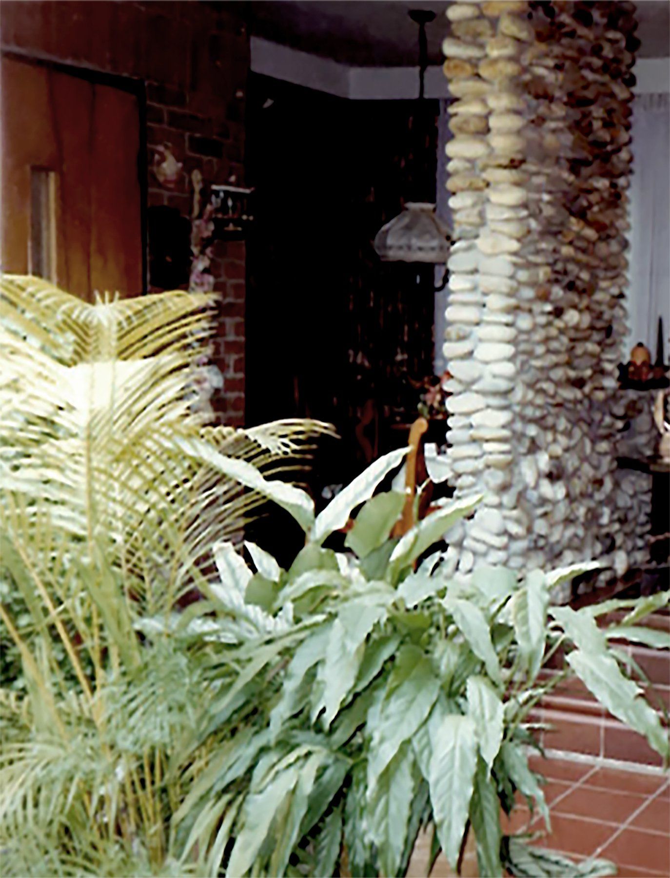 Potted plants in the foreground frame a stone column and a dimly lit indoor space with brick walls and tiled floors.