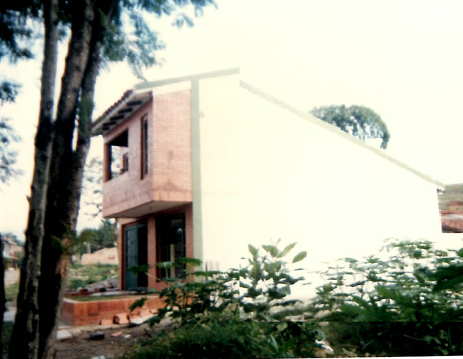 A two-story building with white walls and an exposed brick front section, surrounded by trees and greenery.