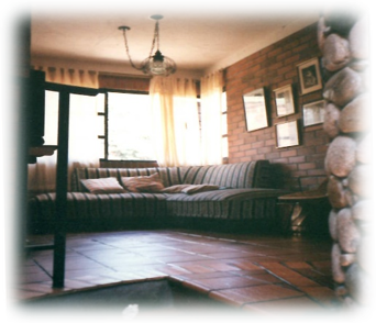 A cozy living room with a corner sofa, brick wall, framed art, tile flooring, and a stone pillar under warm lighting.