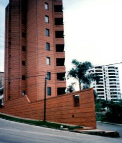 A tall brick apartment building rises above a low, angled brick wall on a sloping street corner, with another tower nearby.