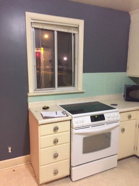 A kitchen corner with dark purple and light green walls, a white stove, cream cabinets, and a window with a blind.