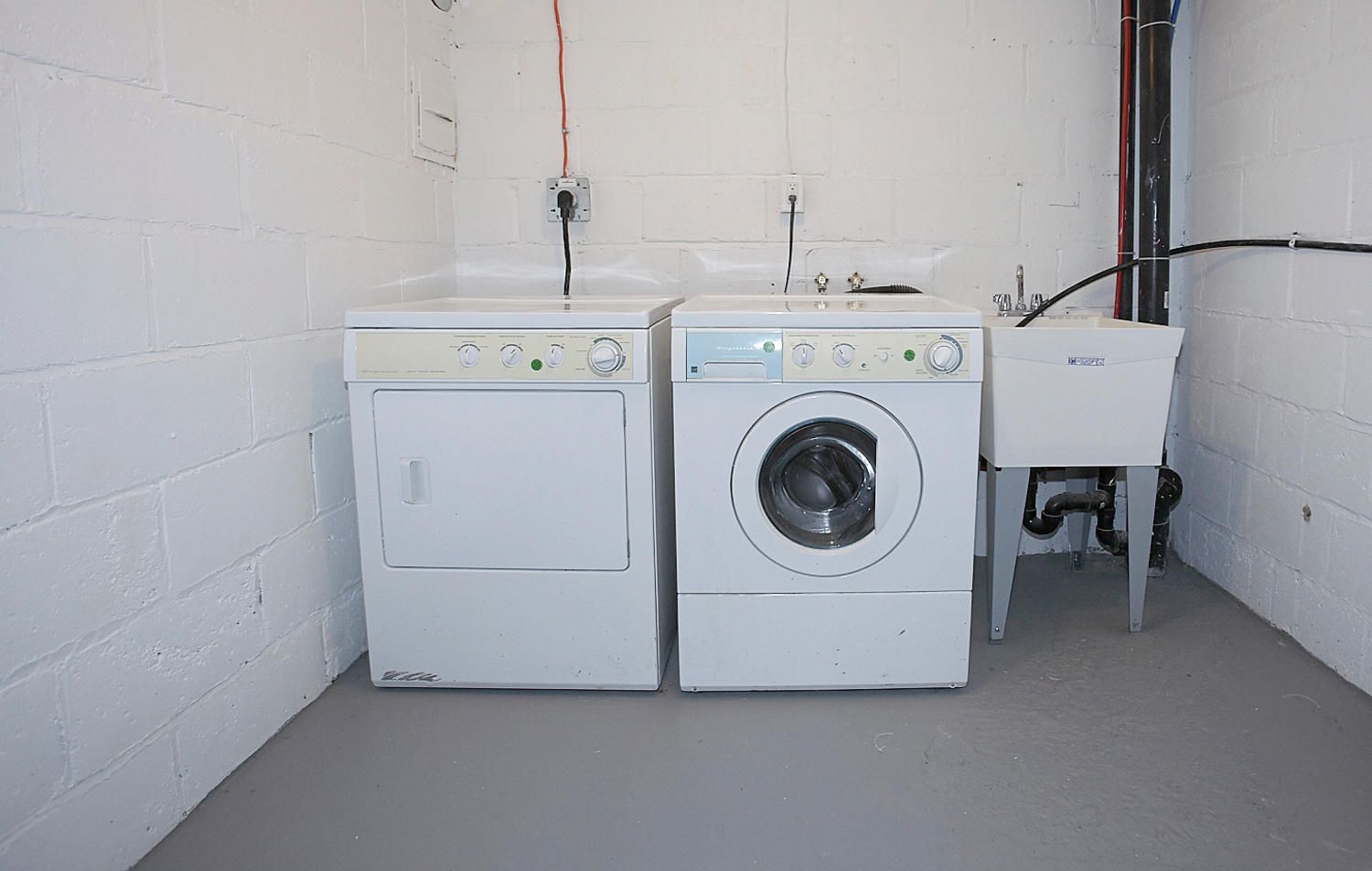 A white dryer and front-loading washer stand next to a utility sink against a white cinder block wall in a laundry room.