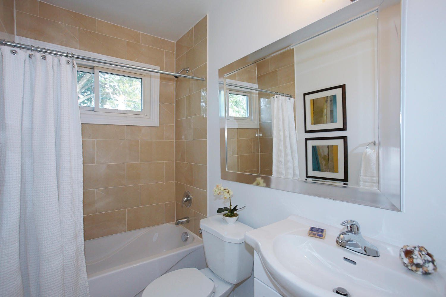 A bright bathroom with a white vanity sink, a toilet, a shower-tub combo with tan tiling, and a large wall mirror.