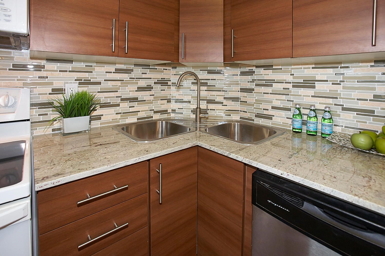 A corner kitchen featuring wooden cabinets, light granite countertops, a tiled backsplash, and a double stainless sink.