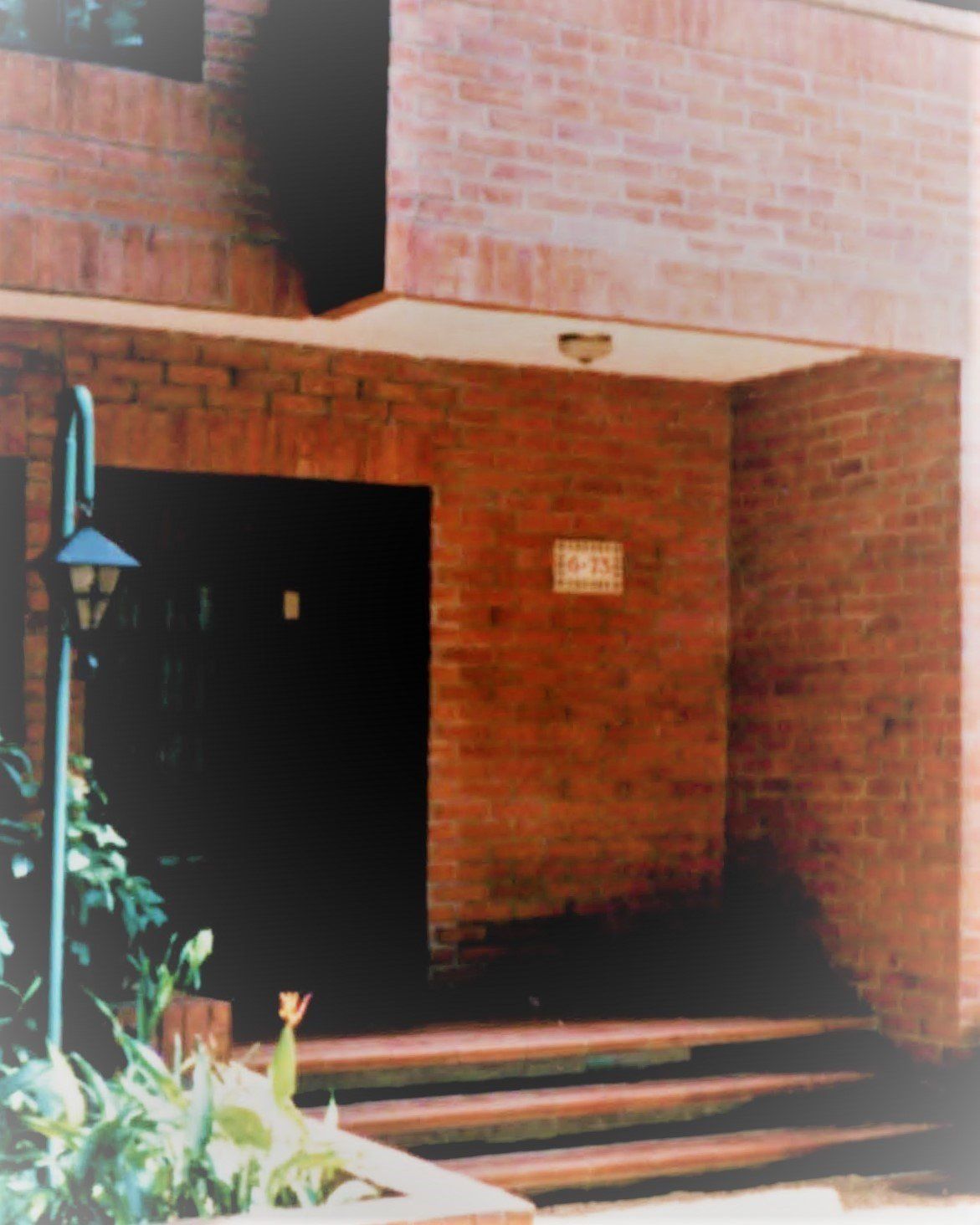 Red brick entryway with a dark door, outdoor lamp, steps leading up, and a small address plaque on the wall.