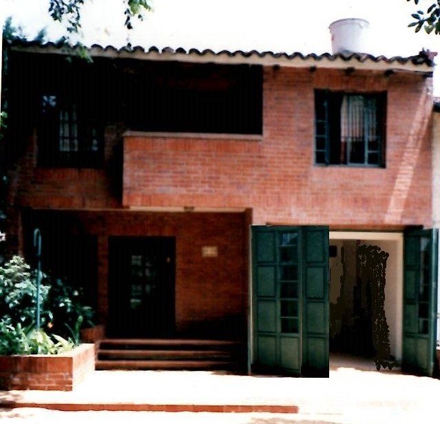 A two-story red brick house with a dark green door, large open green shutters, and a small front garden.