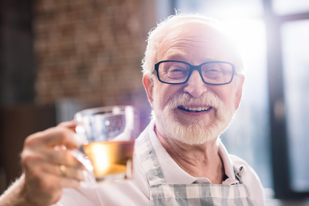 Smiling elderly man with glasses holding a glass of amber liquid, possibly beer, raising it in a toast indoors.