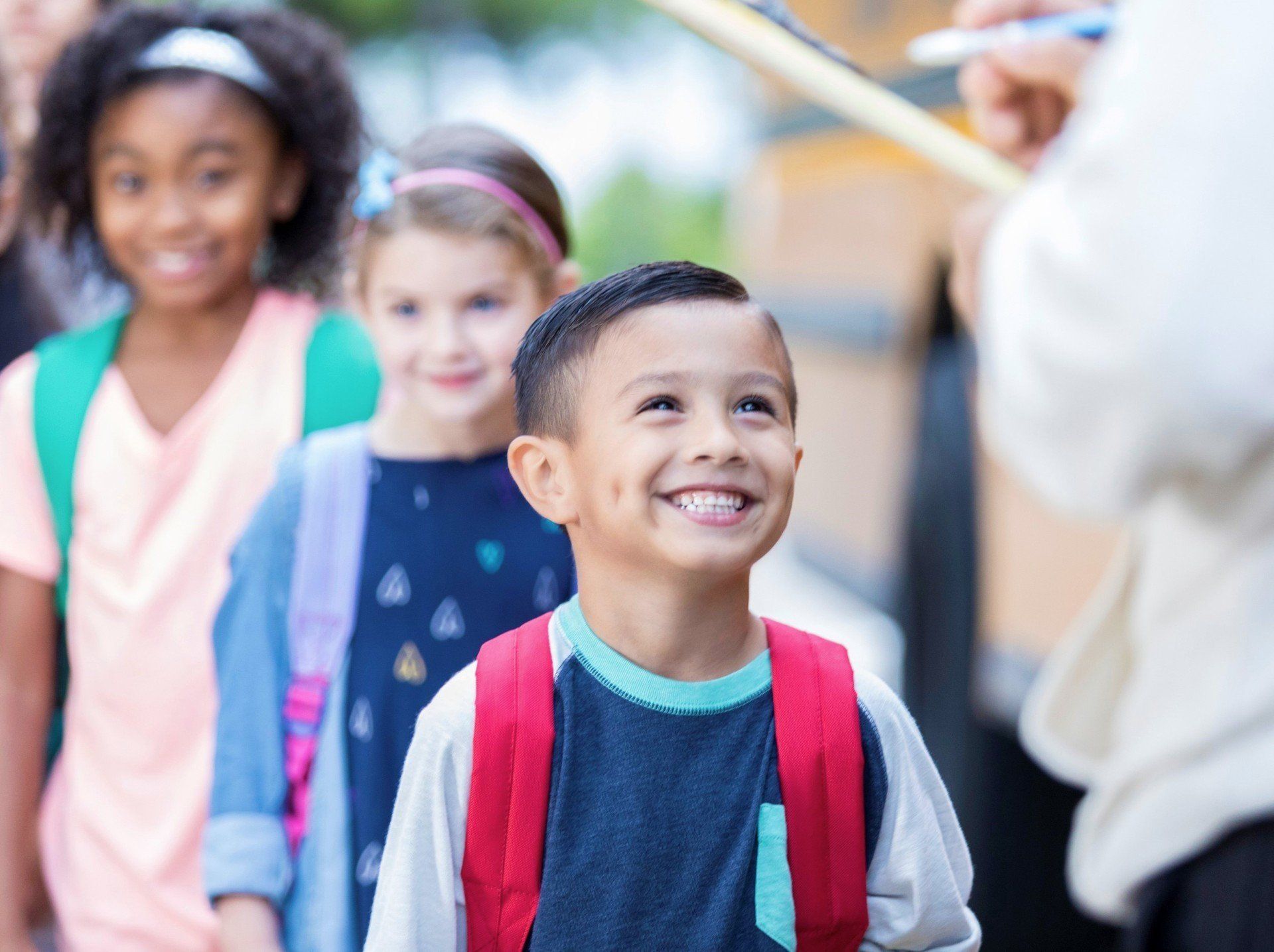 Smiling boy with backpack, standing in line with other children, likely waiting to board a school bus.