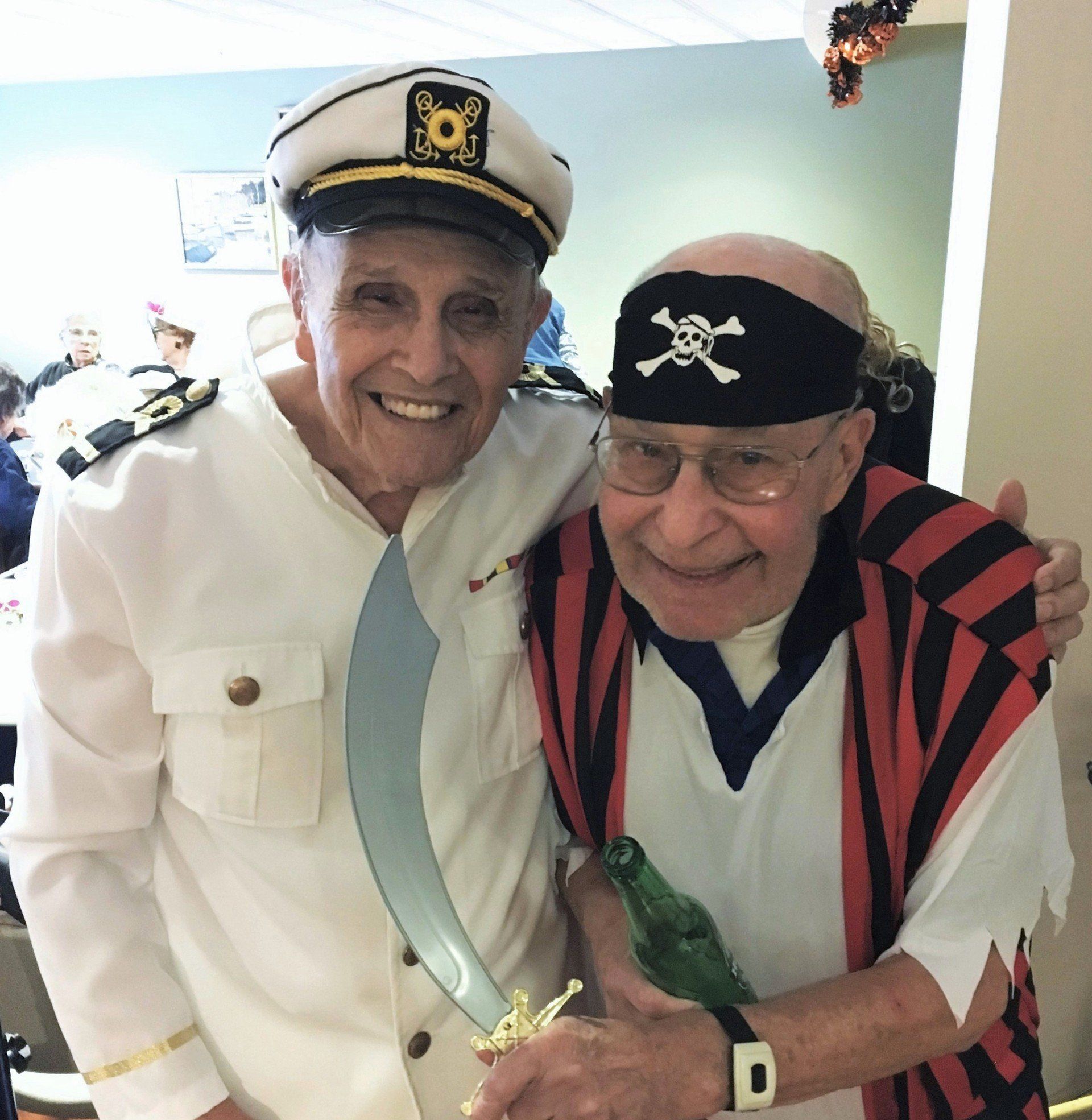 Two older men in costume: One a captain, the other a pirate, arm in arm, smiling at the camera in a festive setting.