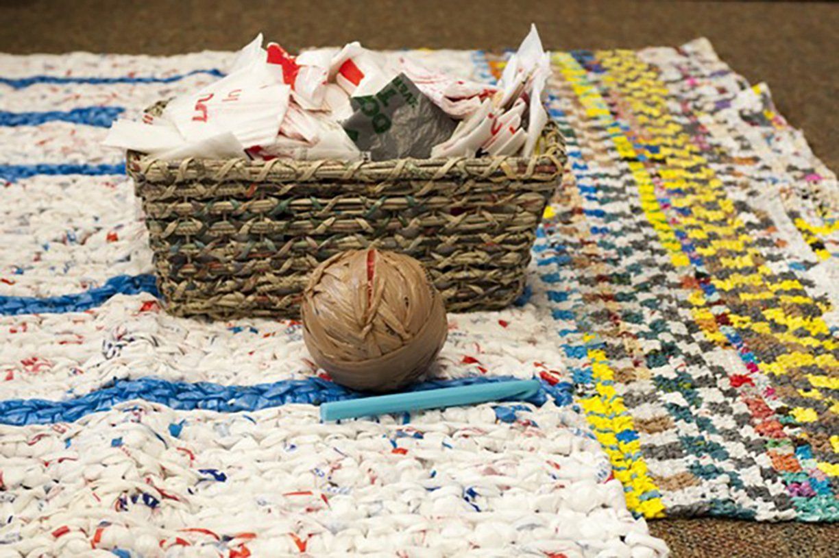 A basket filled with plastic bags sits on a rug made from plastic bags; a ball and pencil are on the rug.