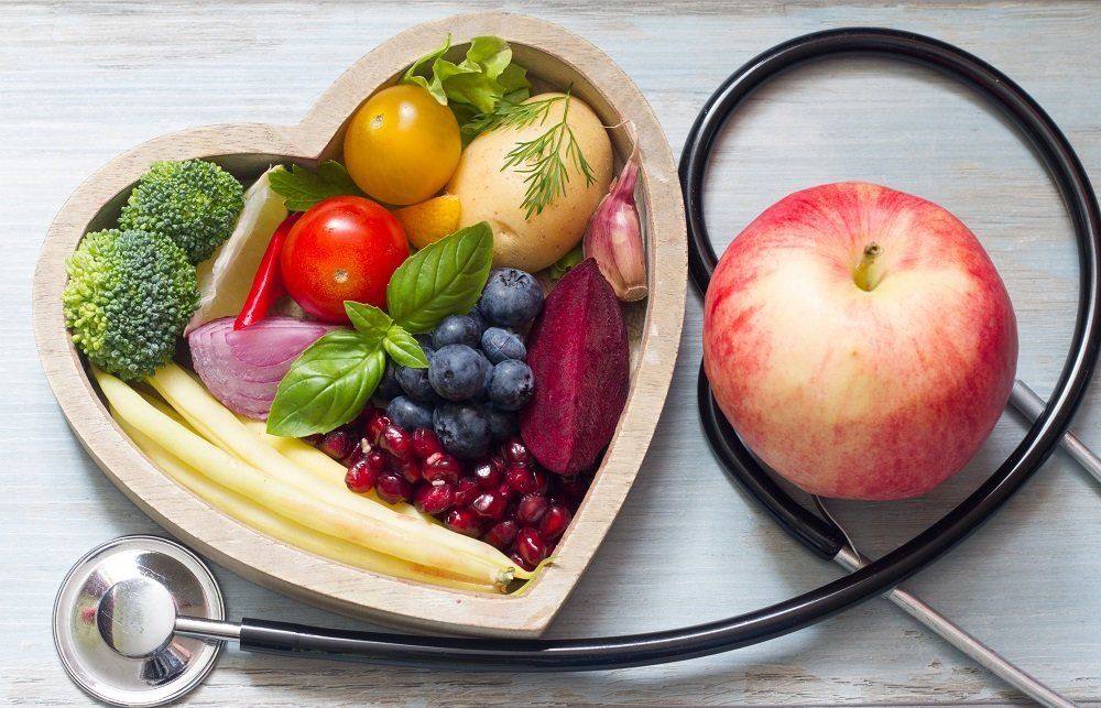 Heart-shaped bowl filled with colorful fruits and vegetables next to a stethoscope and an apple, promoting heart health.