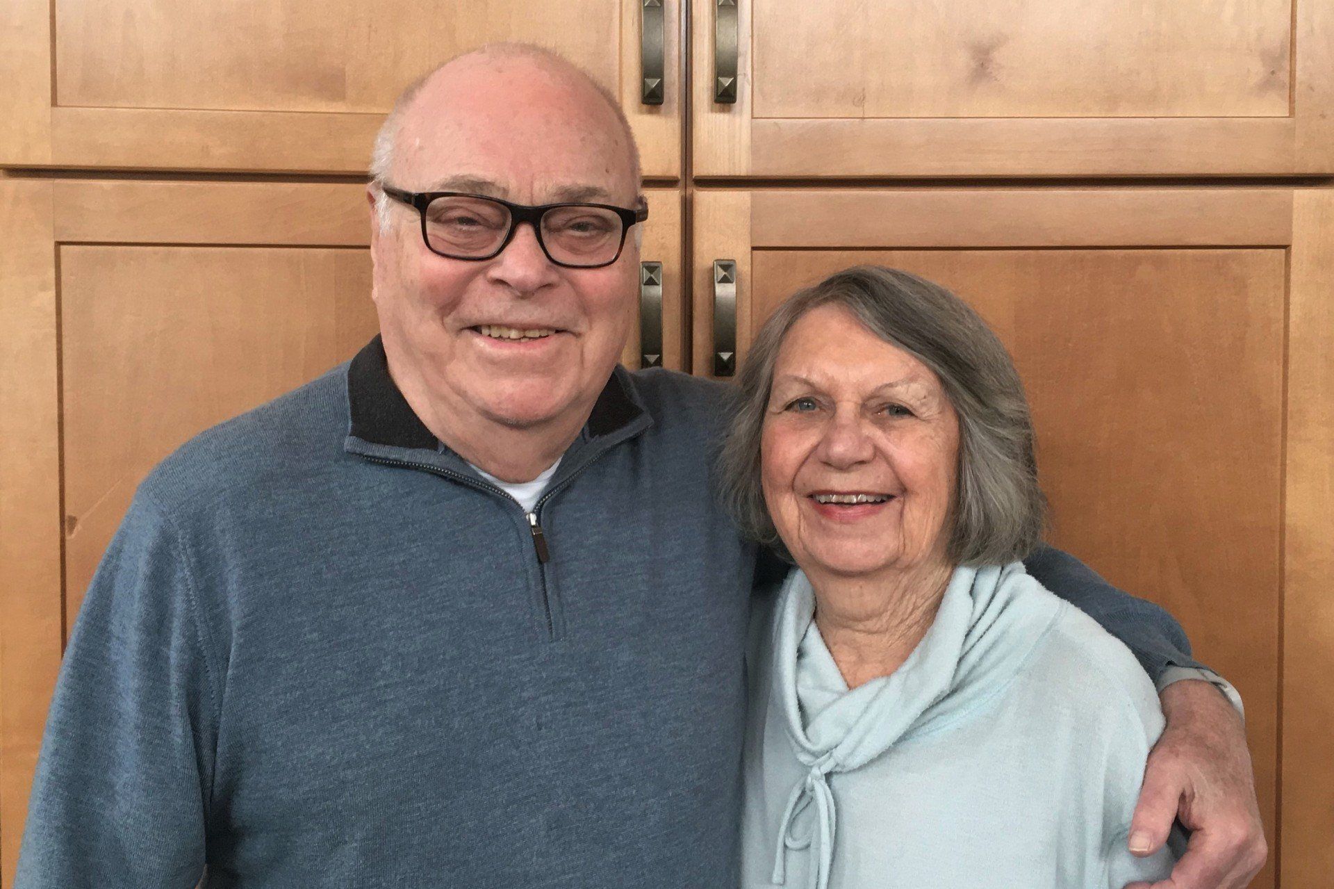 Older couple smiling together, man with glasses and blue sweater, woman with light blue top, in front of wooden cabinets.