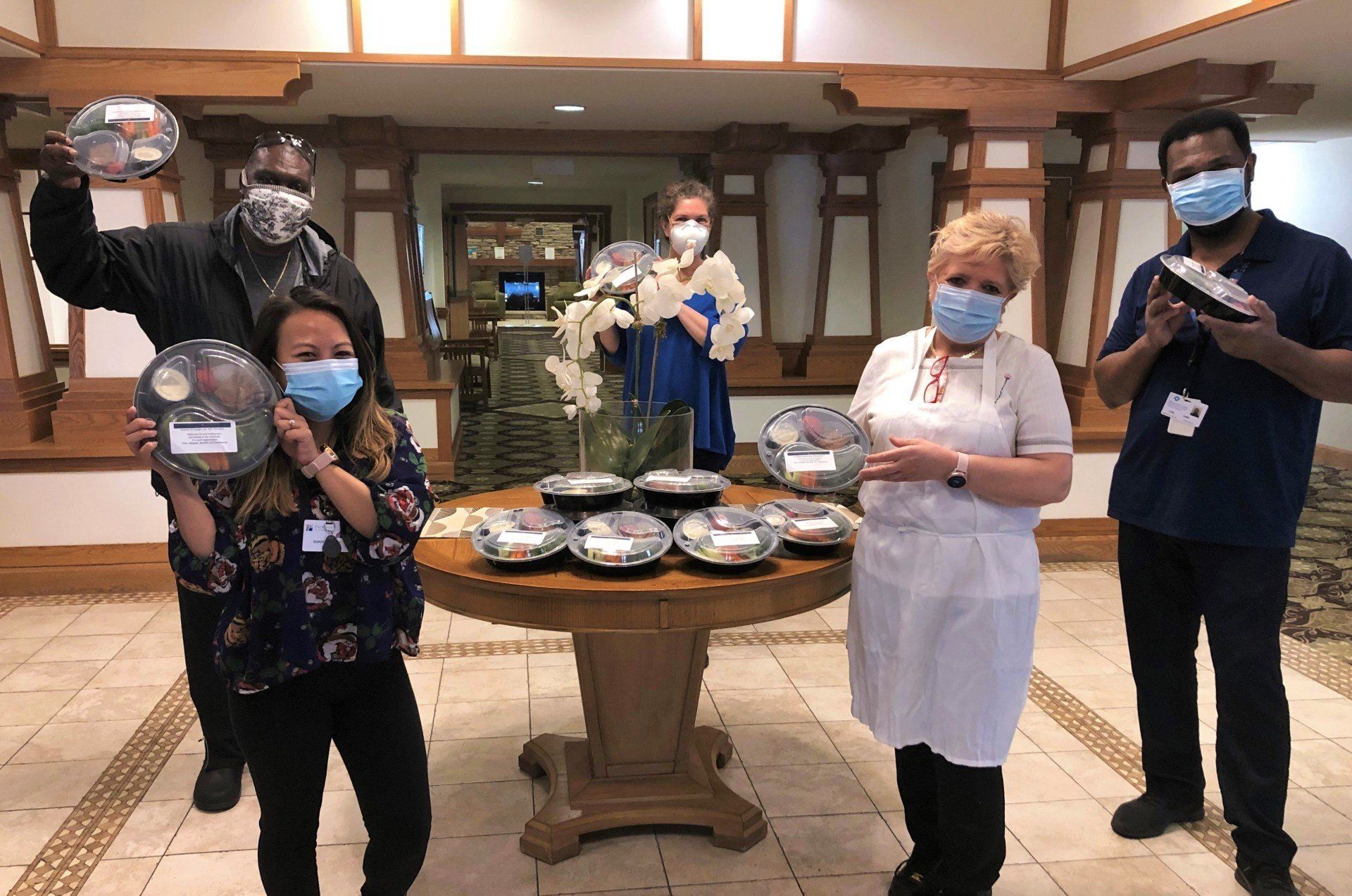 Five masked individuals holding meals in a wood-paneled room. A table in the center displays more meals.