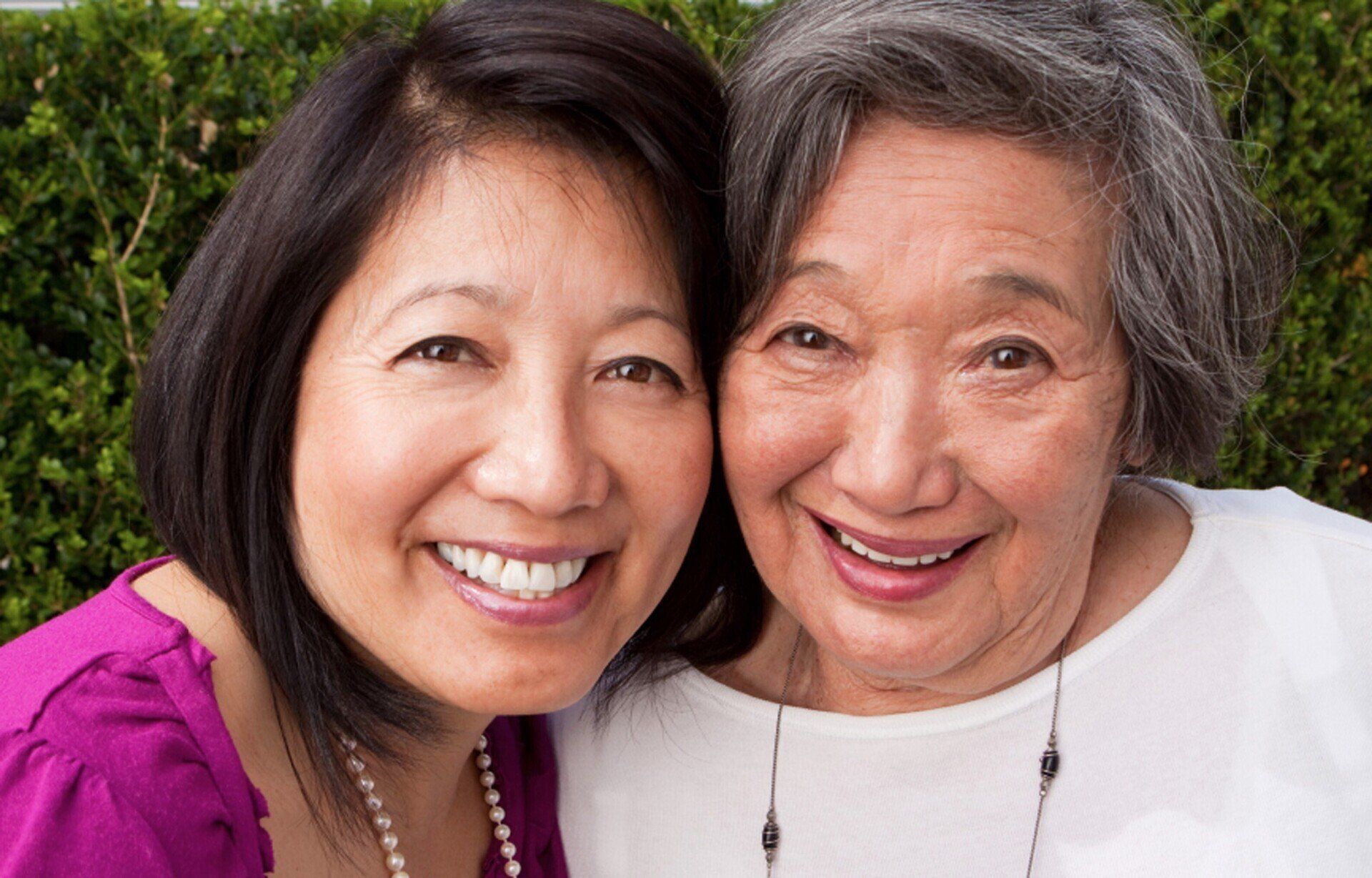 Asian woman and older woman smiling together, close-up portrait, outdoors. The younger woman wears a purple shirt, the older woman a white top.