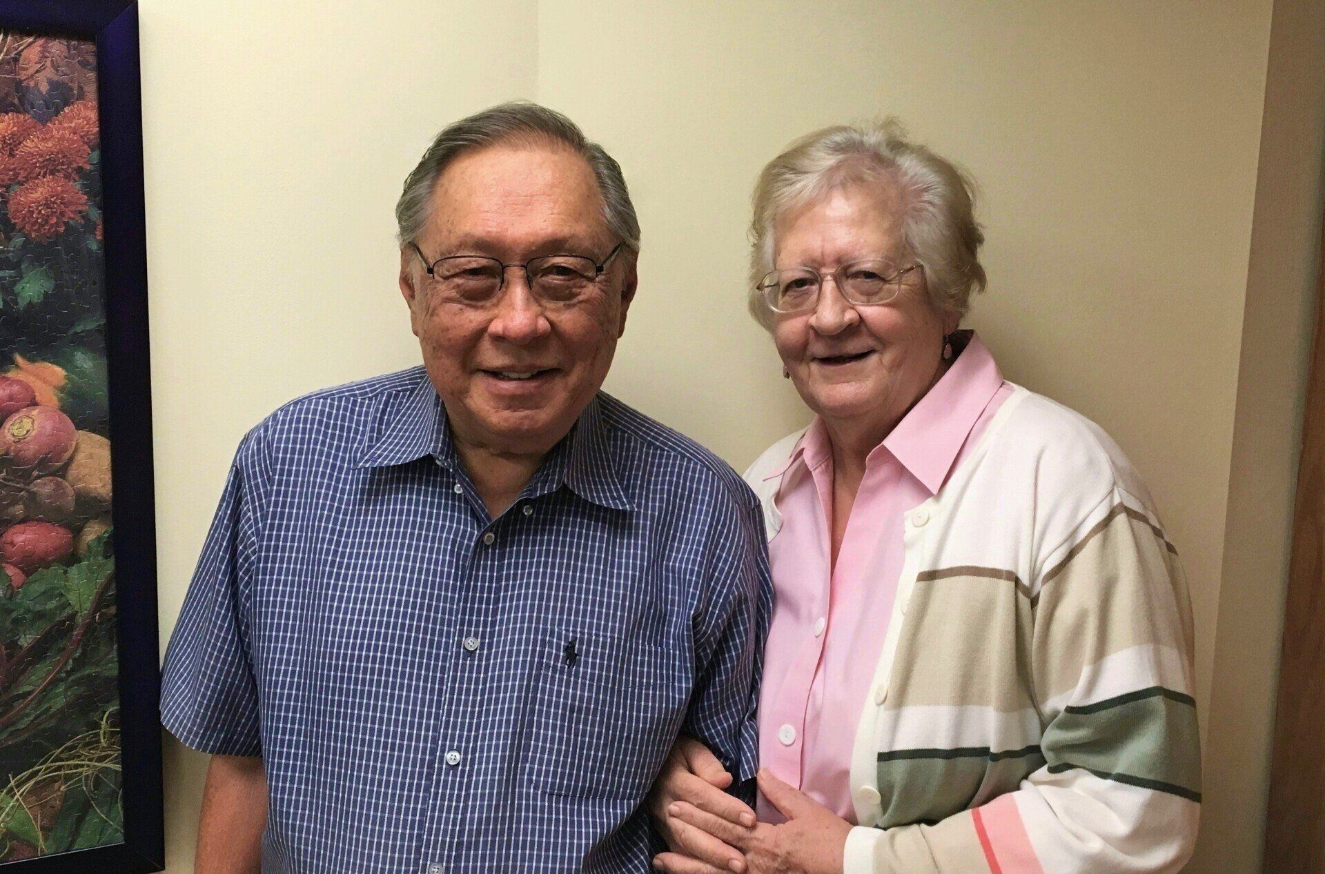 An older Asian man and a Caucasian woman stand close together, smiling. The man wears a blue striped shirt; the woman wears a pink shirt and cardigan.