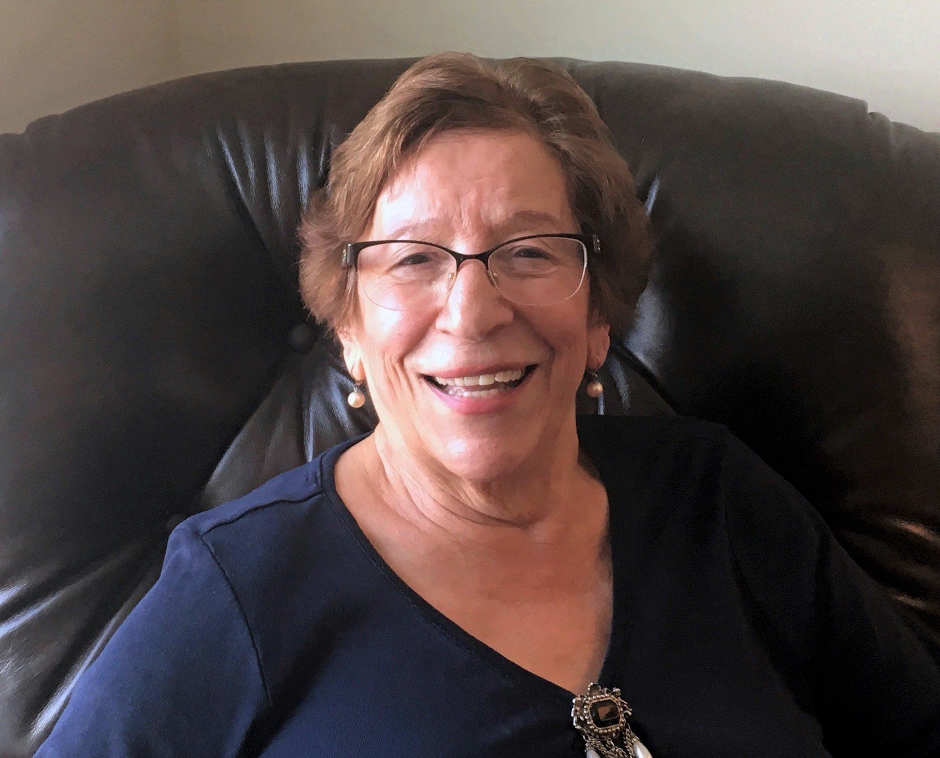 Smiling older woman with glasses sitting in a black chair, wearing a dark blue top.