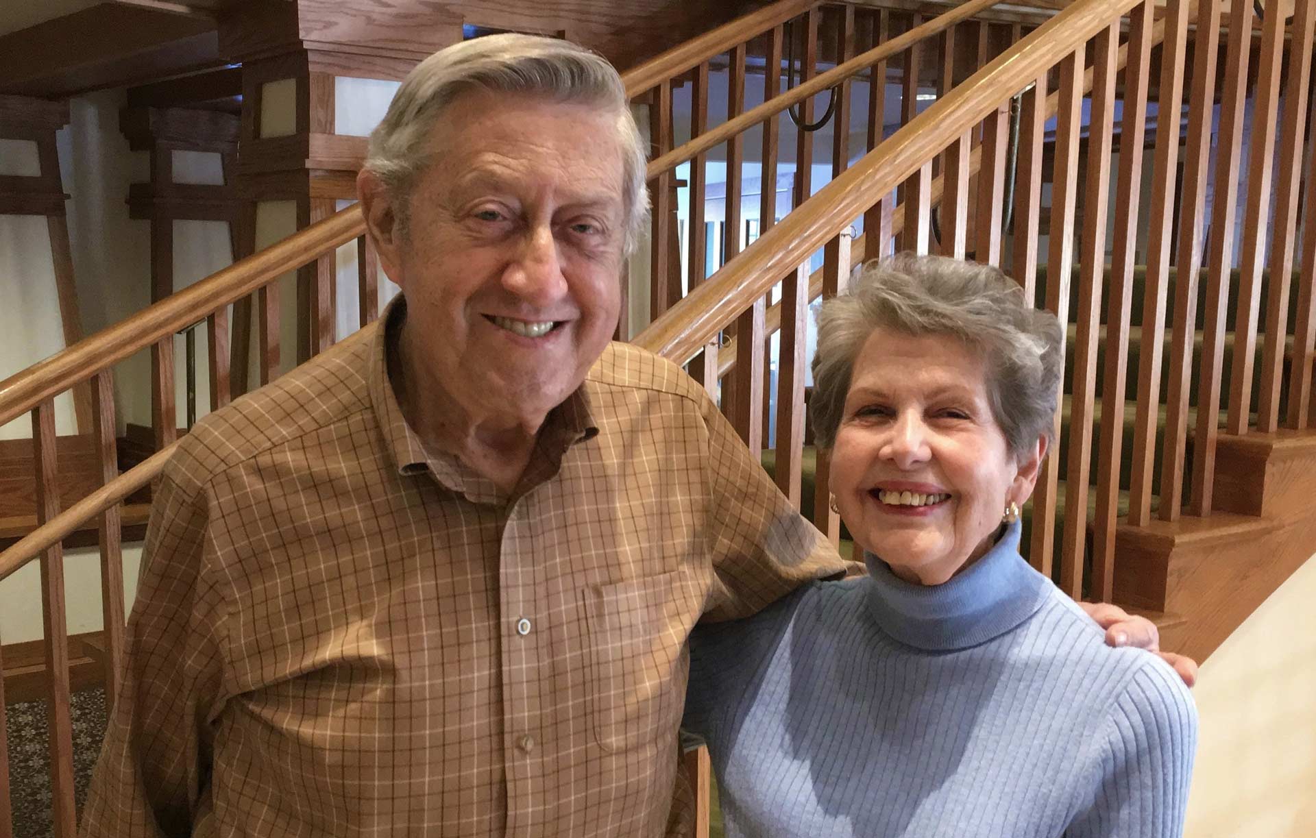 An older smiling couple posing near a wooden staircase. The man wears a brown shirt, and the woman a light blue turtleneck.