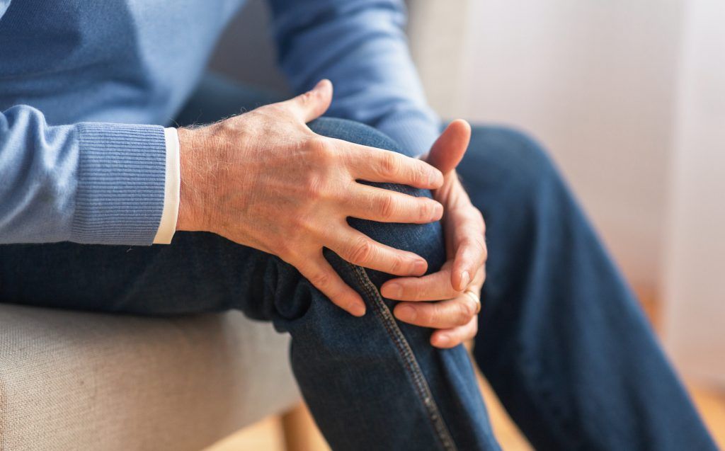 Man holding his painful knee, wearing blue jeans and a sweater, sitting in a chair.