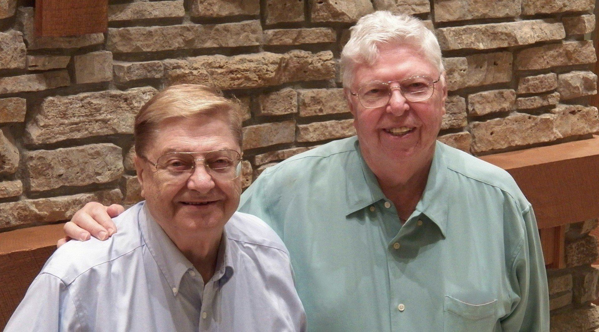 Two older men smiling, standing in front of a stone wall. One man has his arm around the other's shoulder.