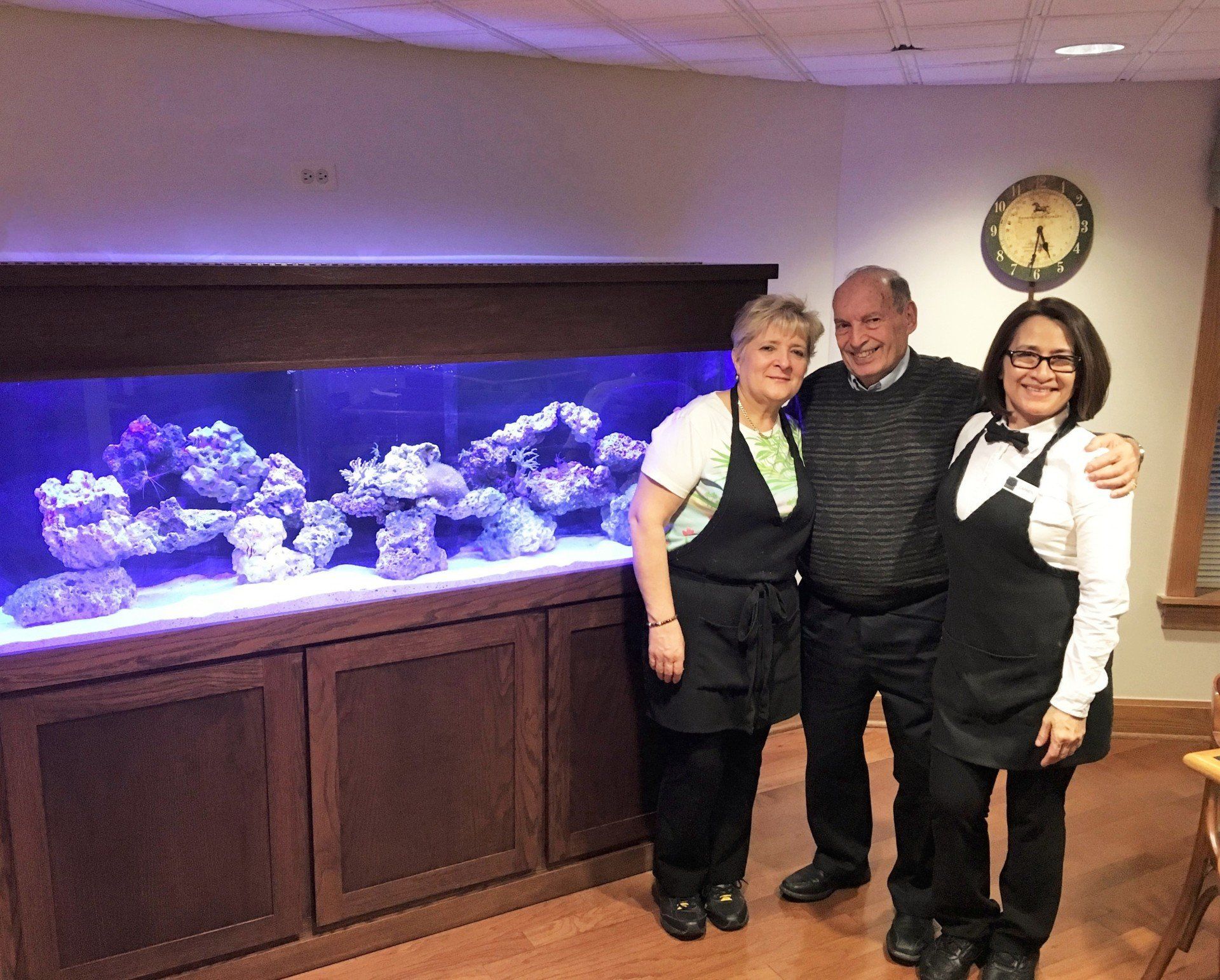 Three people pose in front of a large aquarium; a woman in green smiles, a man in a sweater smiles, and a woman with an apron smiles.