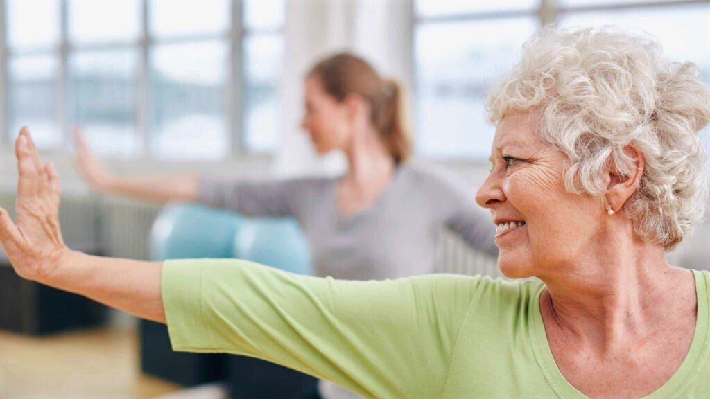 An elderly woman smiles while stretching her arm in a light-filled studio; another woman exercises in the background.