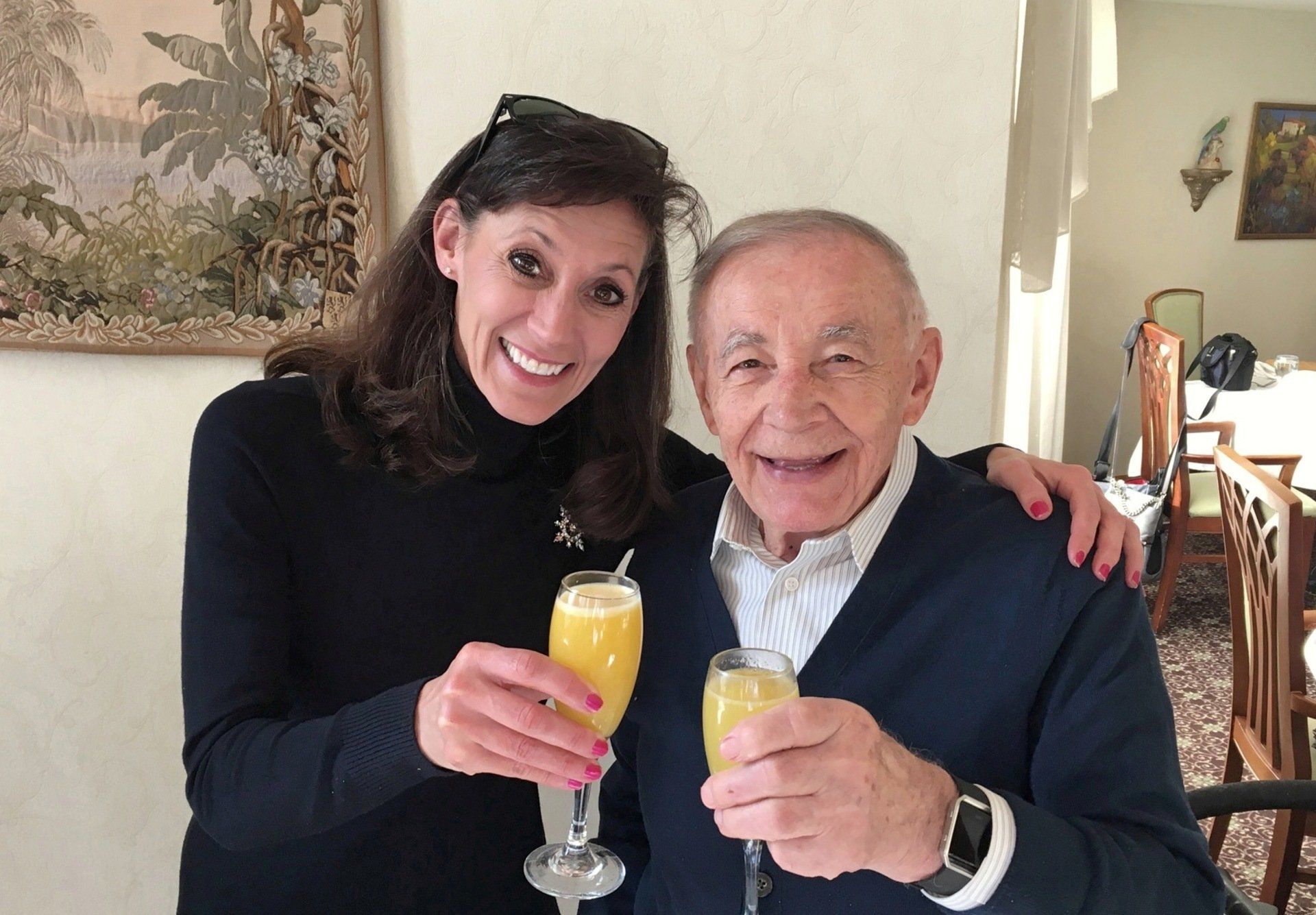 Woman and elderly man smiling and toasting with champagne flutes in a room. The woman has an arm around the man.