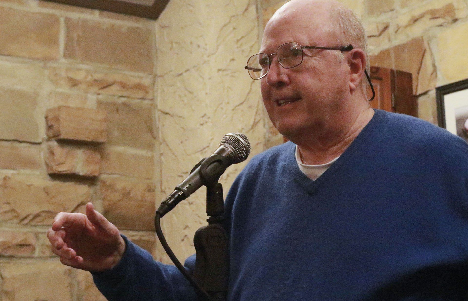 A man with glasses and a blue sweater speaks into a microphone, gesturing with his hand in front of a stone wall.