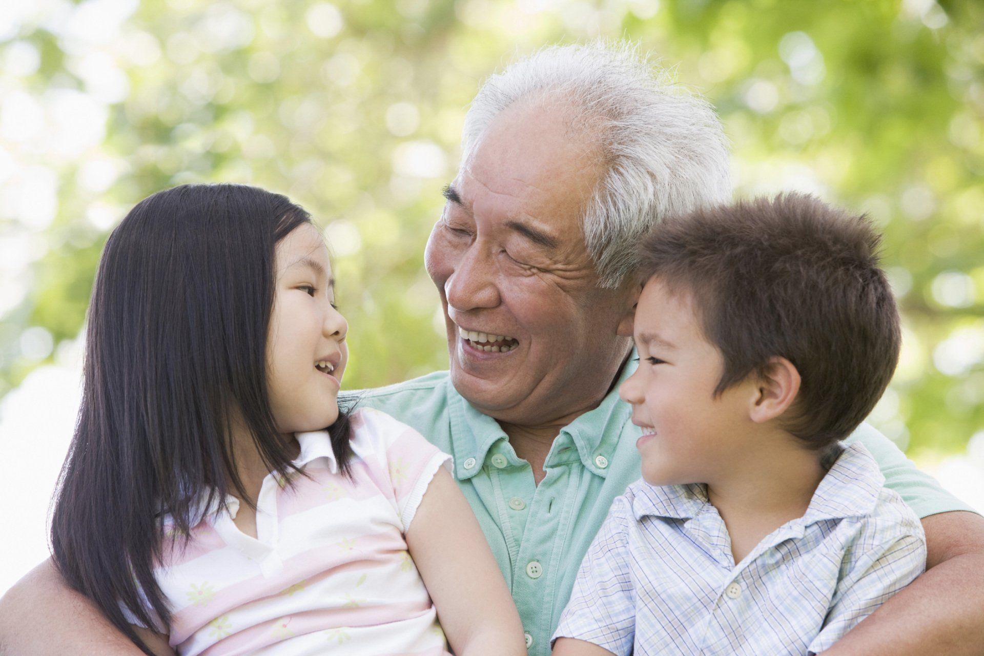 An Asian grandfather with gray hair smiles while holding and looking at two young grandchildren in a sunny outdoor setting.