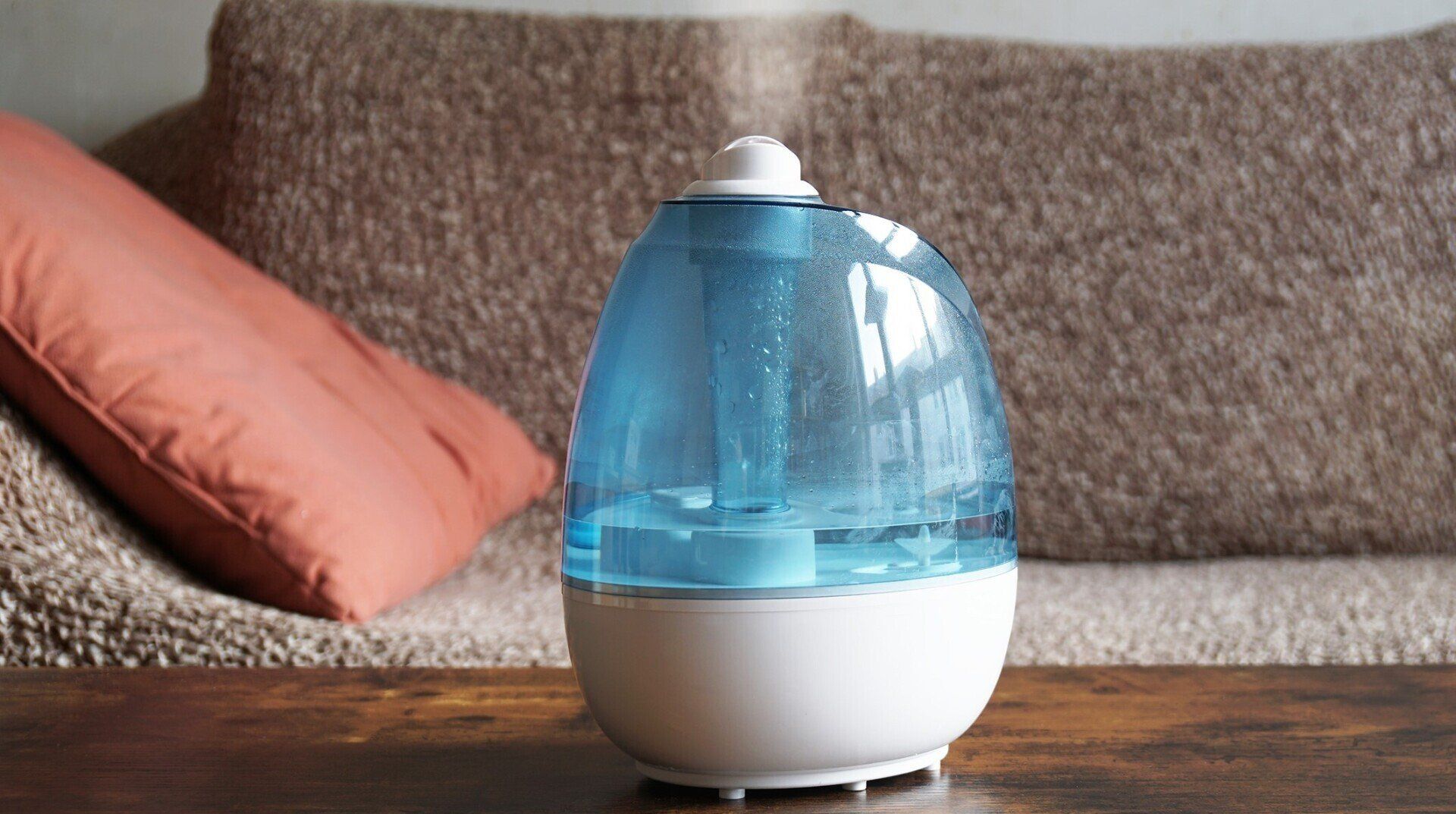 Humidifier on a wooden surface, with a blue water reservoir and white base. An orange pillow and brown couch are in the background.