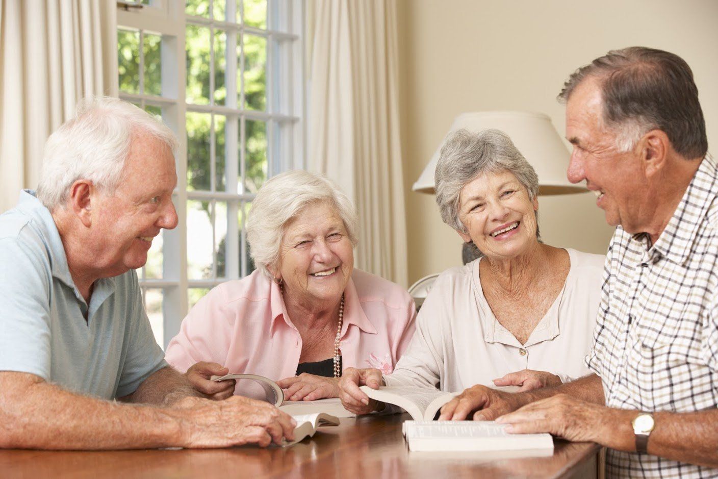 Four older adults seated around a table, smiling and reading a book together in a well-lit room.