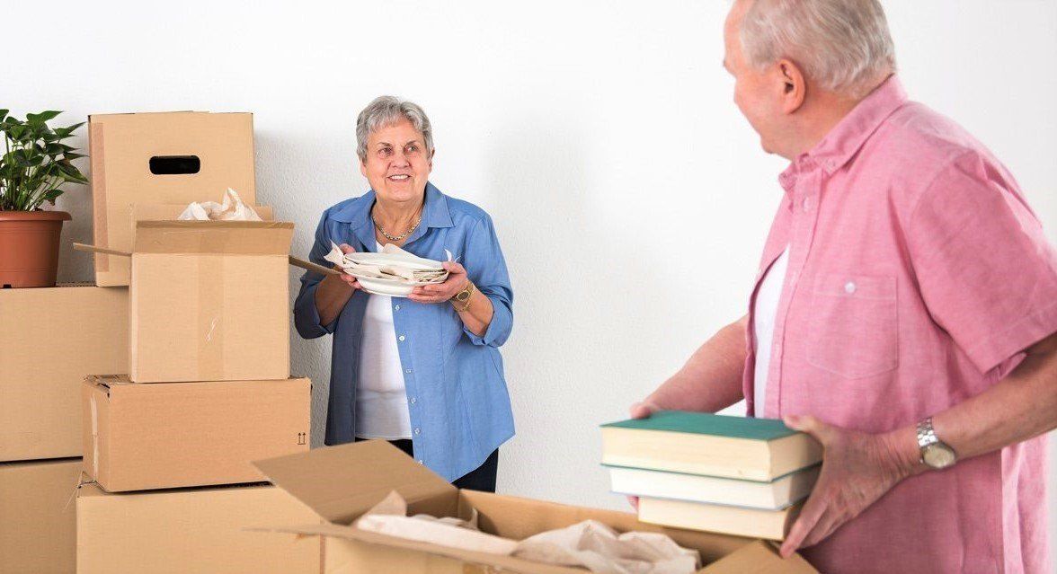 Elderly couple packing moving boxes; woman holding items, smiling; man putting books in a box, also smiling. White wall backdrop.