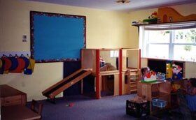 Classroom play area with shelves, toys, a blue bulletin board, and a small ramp under a loft bed