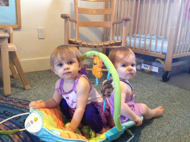 Two toddlers sit on a colorful play mat with a toy arch in a living room.