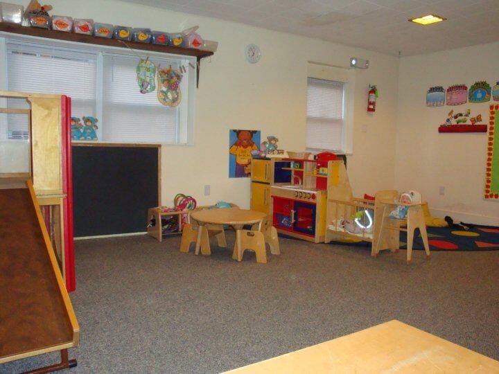 Empty preschool classroom with colorful toys, low tables, chalkboard, and carpeted play area