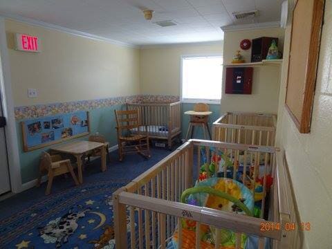 Empty daycare playroom with cribs, toys, chairs, and a blue rug by a bright window.