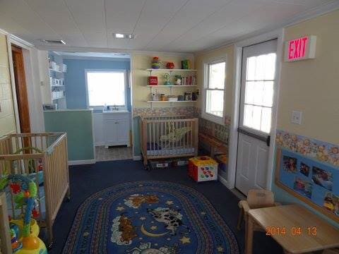 Childcare room with cribs, shelves, toys, and blue carpet, lit by a window and door at the back