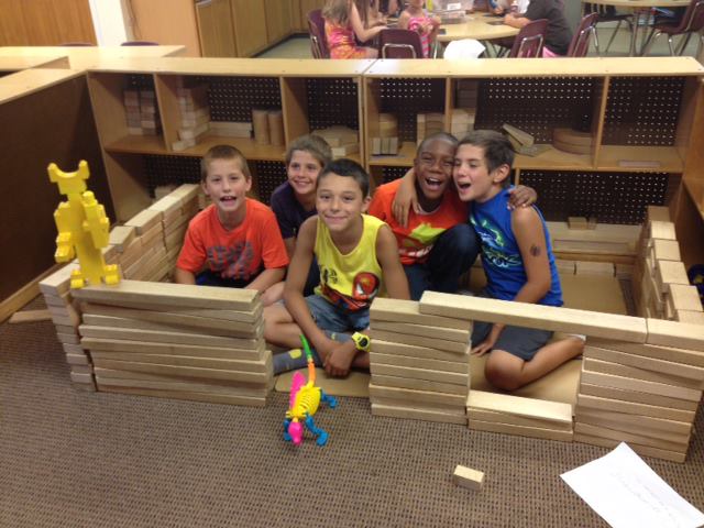 Children inside a block fort in a classroom, smiling beside colorful toys and wooden blocks.