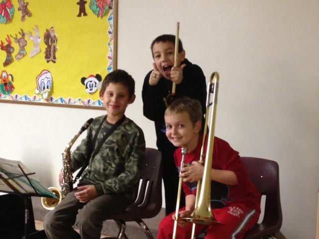 Three children seated with trombones in a classroom, smiling toward the camera.