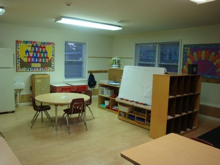 Empty classroom with tables, chairs, shelves, easels, and colorful wall art under fluorescent lights