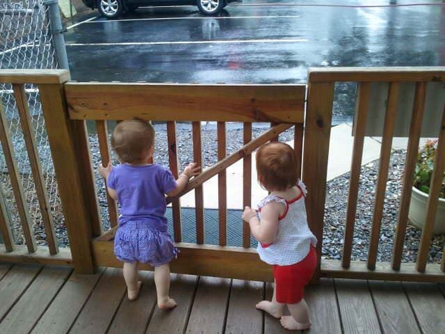 Two small children stand on a porch, looking through a wooden railing toward a wet street.