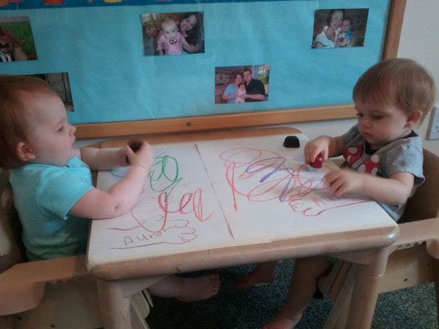 Two toddlers draw with crayons at a small table in a classroom.