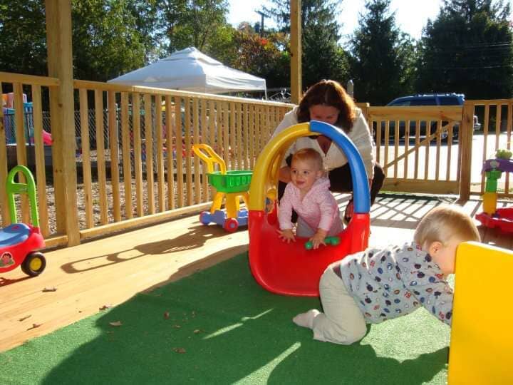 Children playing on a sunny playground deck, one inside a red-and-blue tunnel toy and one crawling nearby.